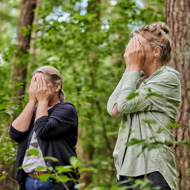 Zwei Personen stehen im Wald und halten sich die Hände vors Gesicht.