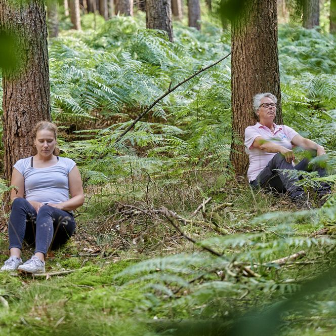 Zwei Frauen lehnen in einem Wald sitzend an einem Baumstamm und haben die Augen dabei geschlossen.