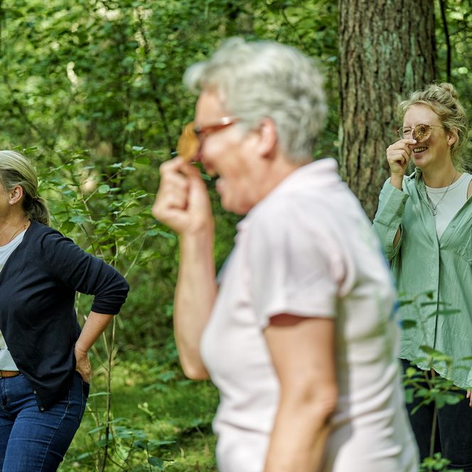 Drei Frauen stehen im Wald und halten sich lachend ein Blatt vors Gesicht.