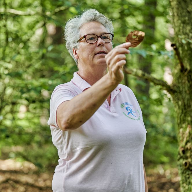Eine Frau steht im grünen Wald und hält ein löchriges Laubblatt hoch.